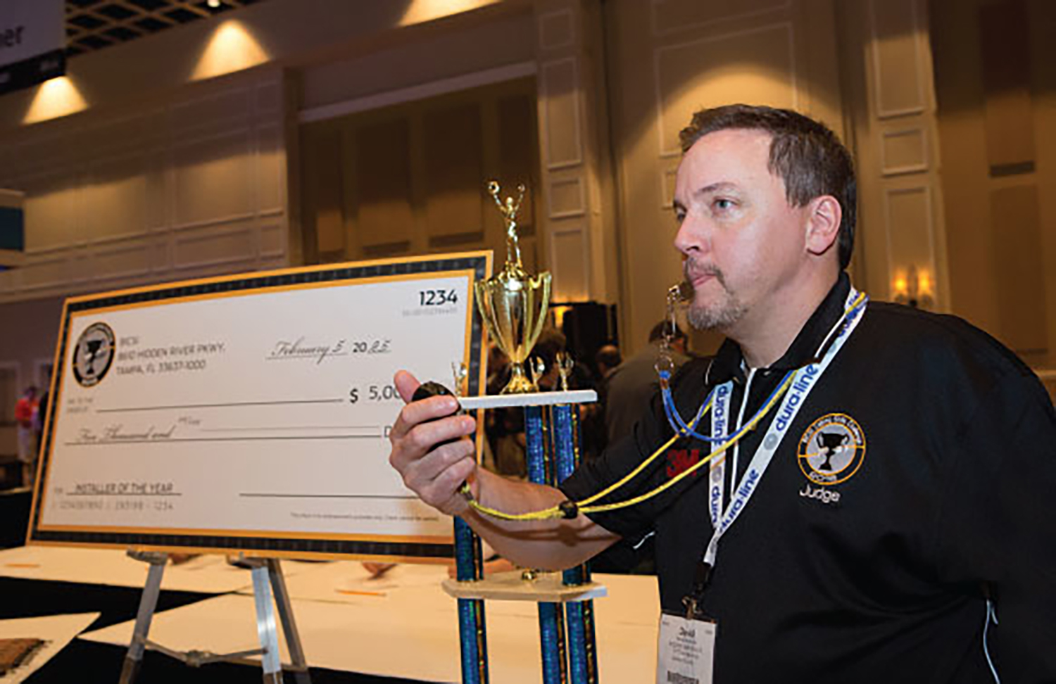 Man in a dark polo shirt stands next to a winner’s trophy and check with a whistle in his mouth and stopwatch in hand