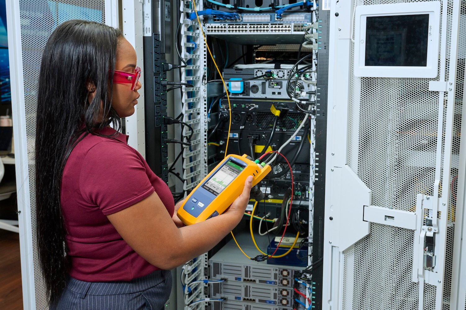 A woman stands in front of a data center server rack while using a fiber cable tester