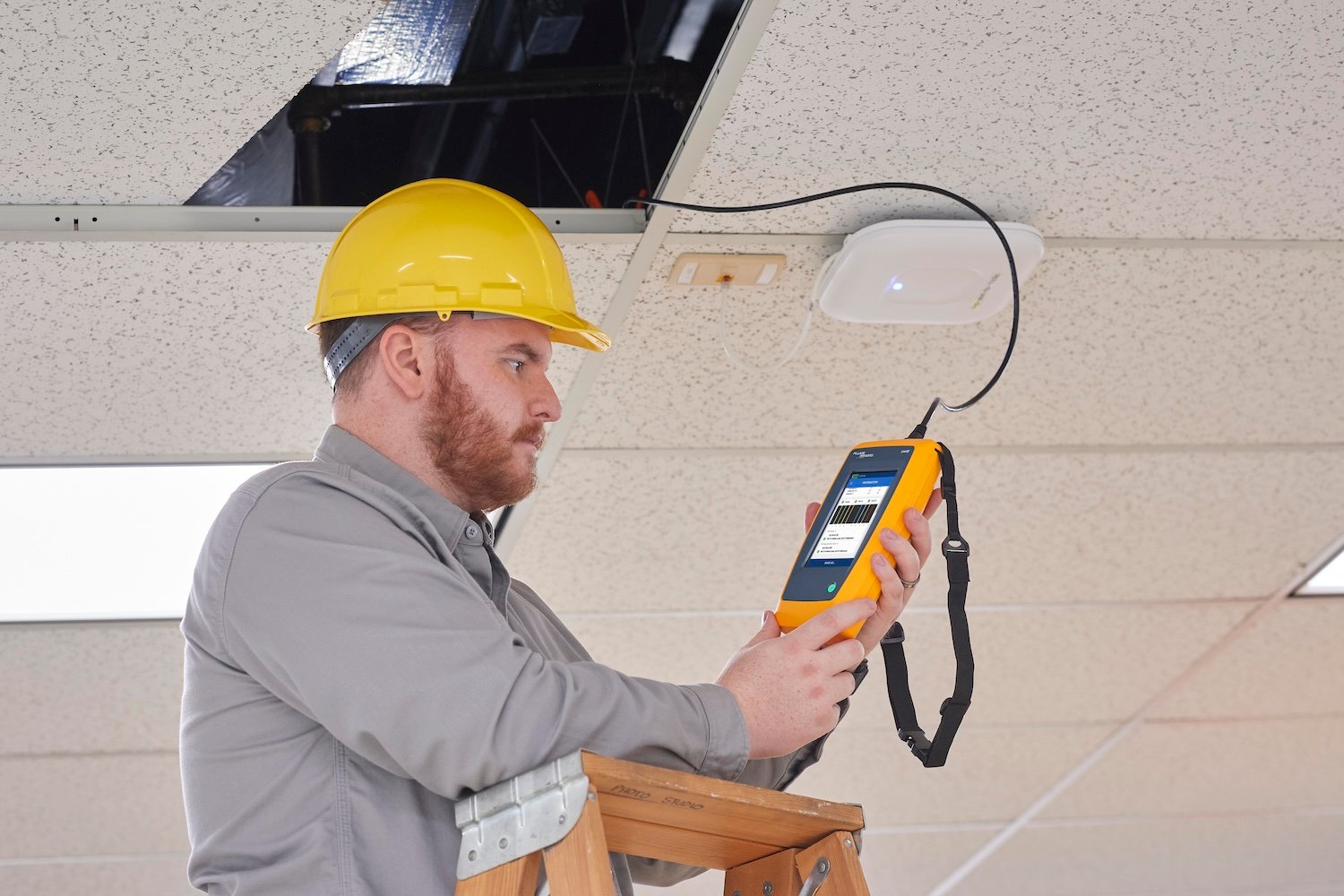 Man in hard hat, standing on a ladder, using a Fluke device under a tile ceiling